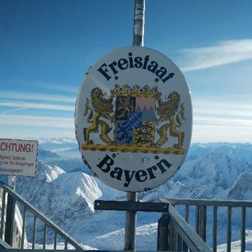 Grenzschild „Freistaat Bayern“ mit dem bayerischen Staatswappen vor einem verschneiten Alpenpanorama
